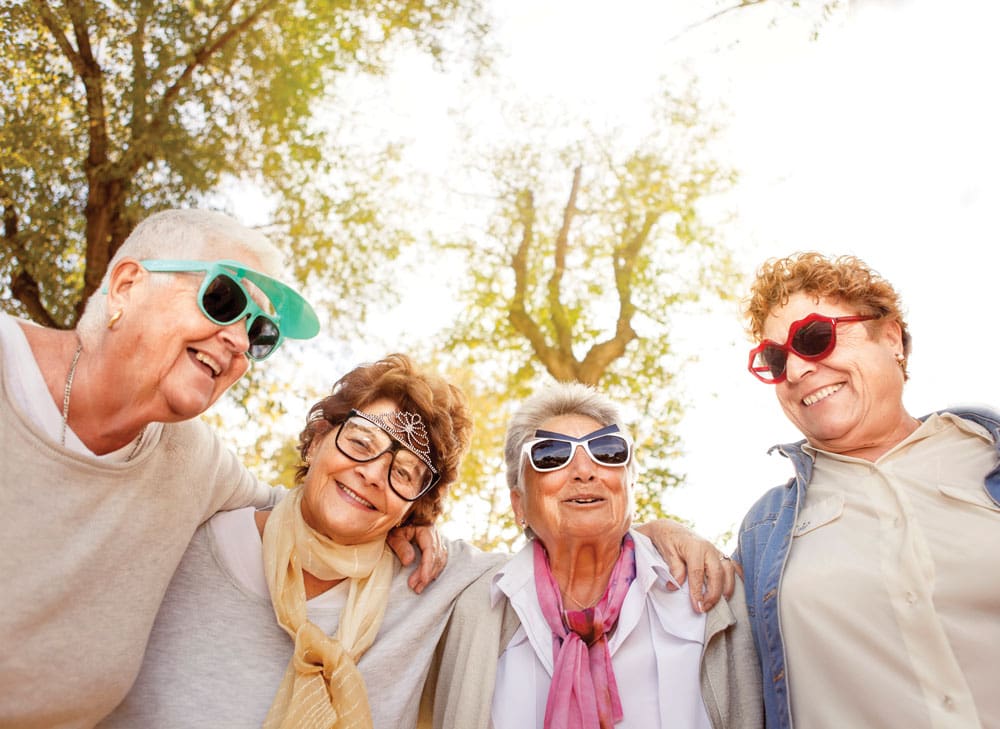 Four older women stand close together outside and pose in funny sunglasses 