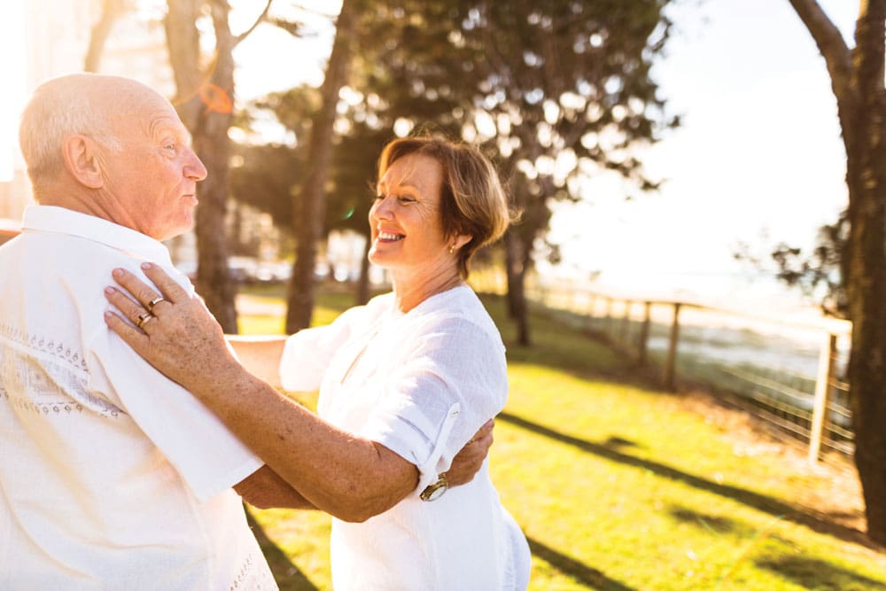 Senior male and female in all white clothing smile and share a dance outside at sunset 
